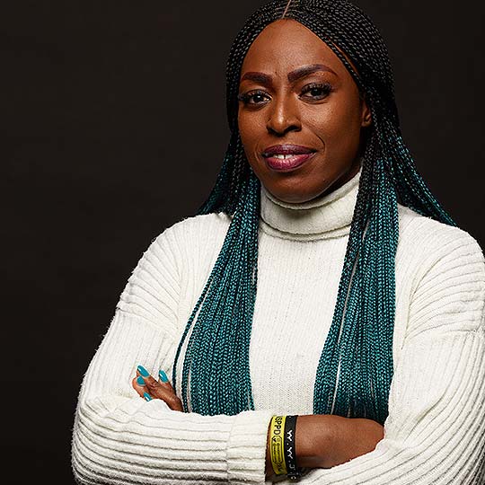 A photograph of Ceewhy stood against a dark background smiling with her arms folded