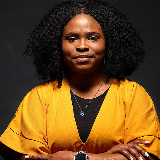 A photograph of Lydia stood against a dark background wearing a mustard-yellow top and smiling